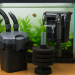 close-up view of three types of aquarium filters—a canister filter, a sponge filter, and a hang-on-back filter—displayed in front of a planted freshwater aquarium with a small fish swimming in the background.