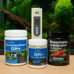 Three GH+ remineralizer products and a digital TDS meter on a wood surface in front of a planted shrimp aquarium with red cherry shrimp